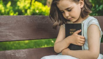 Reading the Holy Bible in outdoors. Christian girl holds bible in her hands sitting on a bench. Concept for faith, spirituality and religion. Peace, hope
